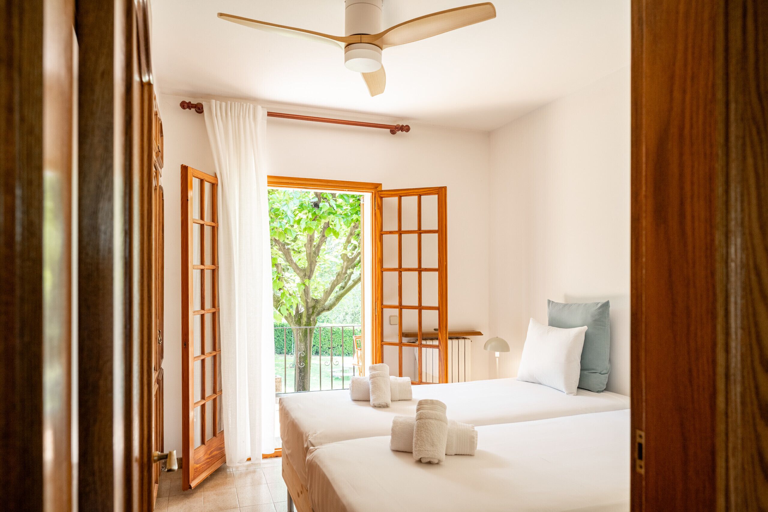A hotel room with two beds, a ceiling fan, a view of greenery outside, and a balcony.