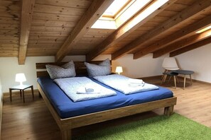 Een slaapkamer met een houten plafond, een bed met blauw beddengoed, kussens, een kleine tafel met een lamp en een stoel.