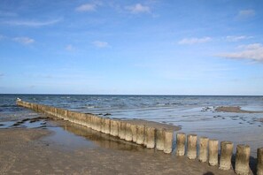 A concrete sea wall with wooden posts, leading to a sandy beach with calm waters. - Holiday home in Warin with swimming pool (Warin)