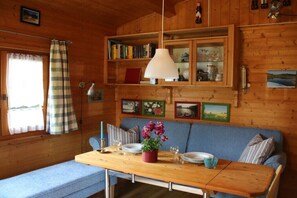 A cabin interior with a wooden dining table, a blue sofa, a bookshelf, and a window with curtains. - Holiday home in Warin with swimming pool (Warin)