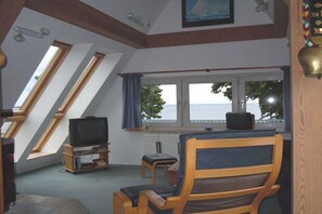 Ein Zimmer mit Fernseher, einem Holzstuhl, einem kleinen Tisch, einem Fenster mit Meerblick und einer schrägen Decke mit Dachfenstern.