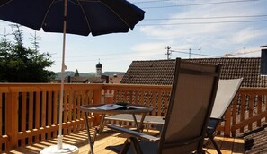 Eine Holzterrasse mit Tisch, Stühlen und einem Sonnenschirm, im Hintergrund sind Gebäude zu sehen.