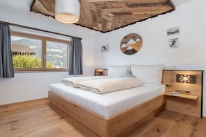 Ein Schlafzimmer mit einem hölzernen Bett, einem Fenster mit Vorhängen, einer Wandleuchte und Blick auf die Berge.