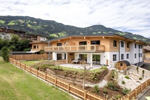 Ein modernes, zweistöckiges Haus mit Holzbalkon, einem eingezäunten Garten und einer Bergkulisse im Hintergrund.