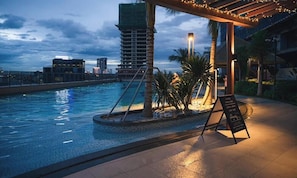 A pool area with a pergola, palm trees, and a view of a city skyline at dusk. - Itadaki Suite (Phnom Penh)