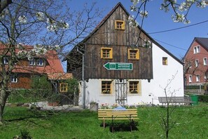 Ein traditionelles Holzgebäude mit weißer Fassade und gelben Fensterrahmen, eine Bank im Vordergrund und ein Wegweiser.