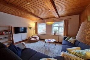 Ein gemütliches Wohnzimmer mit Holzbalkendecke, einem Flachbildfernseher, einer Couch, einem Sitzsack, einem kleinen Tisch und einem Fenster mit Vorhängen.