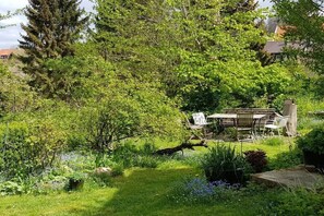 Ein Garten mit einer Holzbank und Stühlen, umgeben von üppigem Grün und Bäumen.