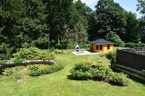 Eine kleine Holzhütte mit einem Reetdach, umgeben von einem gepflegten Garten mit Teich und verschiedenen Sträuchern.