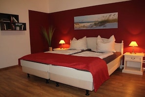 Ein Bett mit roter Decke, zwei Nachttische mit Lampen, ein Bücherregal und ein Bild an der Wand.