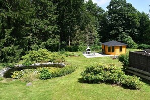 Eine kleine Holzhütte mit Terrasse, umgeben von einem üppigen Garten und einem Teich.