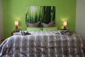 Ein Schlafzimmer mit grüner Wand, einem großen Wandteppich mit Waldmotiv, einer flauschigen grauen Tagesdecke, zwei Nachttischen mit Lampen und zwei Kissen.