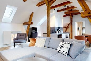 Ein modernes Loft mit freiliegenden Holzbalken, einem grauen Ecksofa, einem Essbereich und einer Küche im Hintergrund.