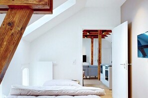 Quarto moderno estilo loft com teto inclinado, estrutura de vigas de madeira, cama com roupa de cama branca e vista para a área de estar adjacente.