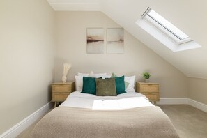 Een slaapkamer met een schuine kap, dakraam, een bed met wit beddengoed, twee nachtkastjes met decoratieve objecten, en twee abstracte schilderijen aan de muur.