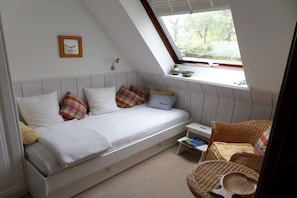 Ein Schlafzimmer im Loft-Stil mit einem Bett, einem kleinen Nachttisch, einem Korbstuhl und einem Fenster mit Blick ins Grüne.