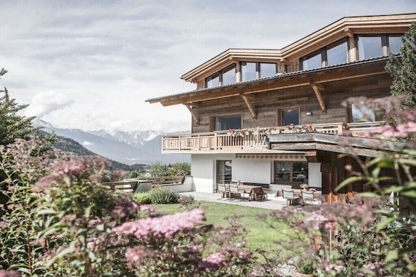 A wooden chalet with a balcony, a patio with outdoor seating, and a mountainous backdrop.