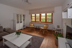 Ein Wohnzimmer mit Sofa, Couchtisch, Korbstühlen, Bücherregal, Heizkörper und einem Fenster mit Jalousien.