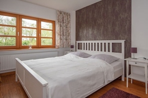 Ein Schlafzimmer mit einem weißen Bett, einem Fenster mit Vorhängen, einem Heizkörper und einem Nachttisch mit Lampe.