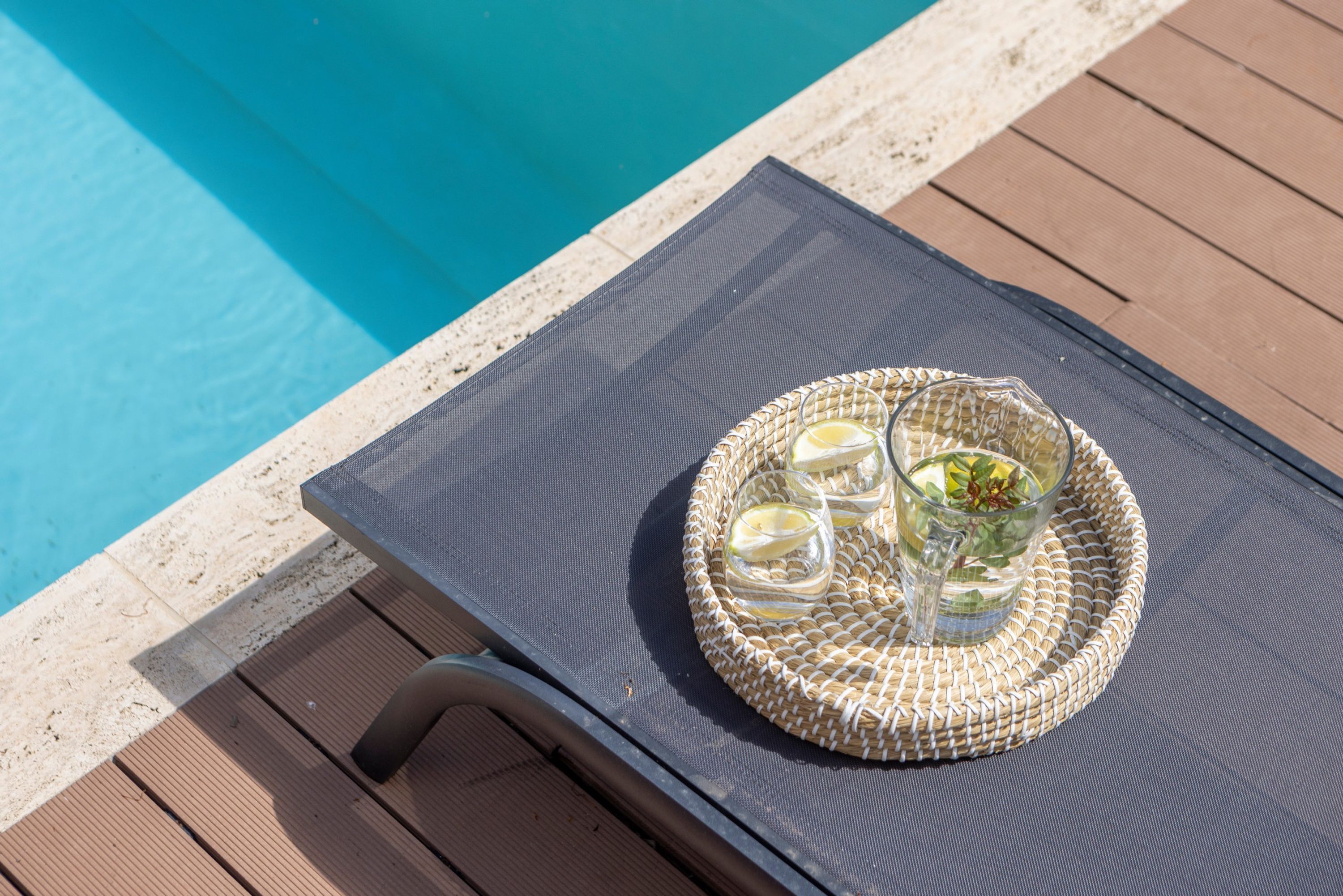 A poolside lounge chair with a woven tray table holding a glass of water, a glass of lemon, and a glass of iced tea.