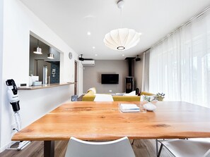 Ett modernt vardagsrum med ett matbord i trä, en soffa, en tv som är monterad på väggen och ett stort fönster med gardiner.