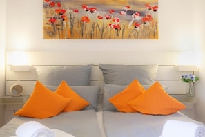Ein Doppelbett mit grauer Bettwäsche, zwei orangefarbene Kissen und ein Gemälde mit roten Blumen an der Wand.