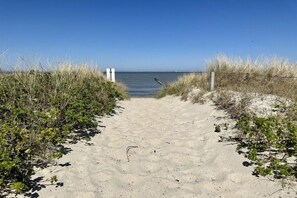 Ein Sandweg am Strand führt zum Meer, ges äumt von Vegetation und einem weißen Gebäude.