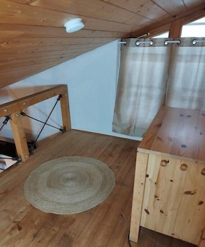 Eine Holzbalkendecke mit einer schlichten Leuchte, ein Holztisch, ein gewebter Teppich und ein Fenster mit Vorhängen.