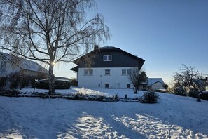 A two-story house with a dark roof and white walls, surrounded by snow-covered ground and leafless trees. - Holiday apartment with 1 bedroom (Altenkirchen Lahn-Dill-Kreis)