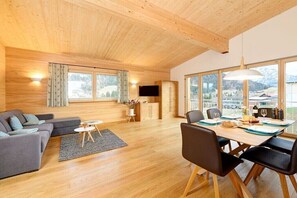 Ein geräumiges Wohnzimmer mit Holzboden, einer Couch, einem Esstisch mit Stühlen und großen Fenstern mit Blick ins Freie.