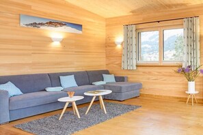 Ein modernes Wohnzimmer mit einem grauen Sofa, zwei kleinen runden Tischen, einer Holzbalkendecke und einem Fenster mit Vorhängen.