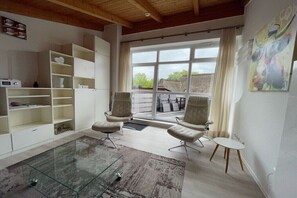 Ein modernes Wohnzimmer mit Glastisch, weißen Regalen, zwei Ledersesseln und einem großen Fenster mit Aussicht.