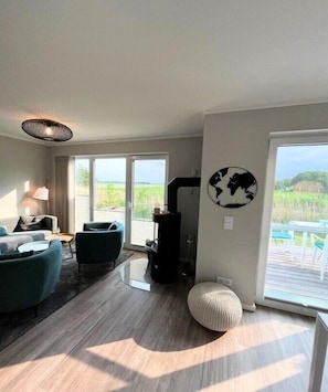 Ein modernes Wohnzimmer mit einer Couch, Sesseln, einem runden Hocker, einem Kamin und einem großen Fenster mit Blick auf Grünflächen.