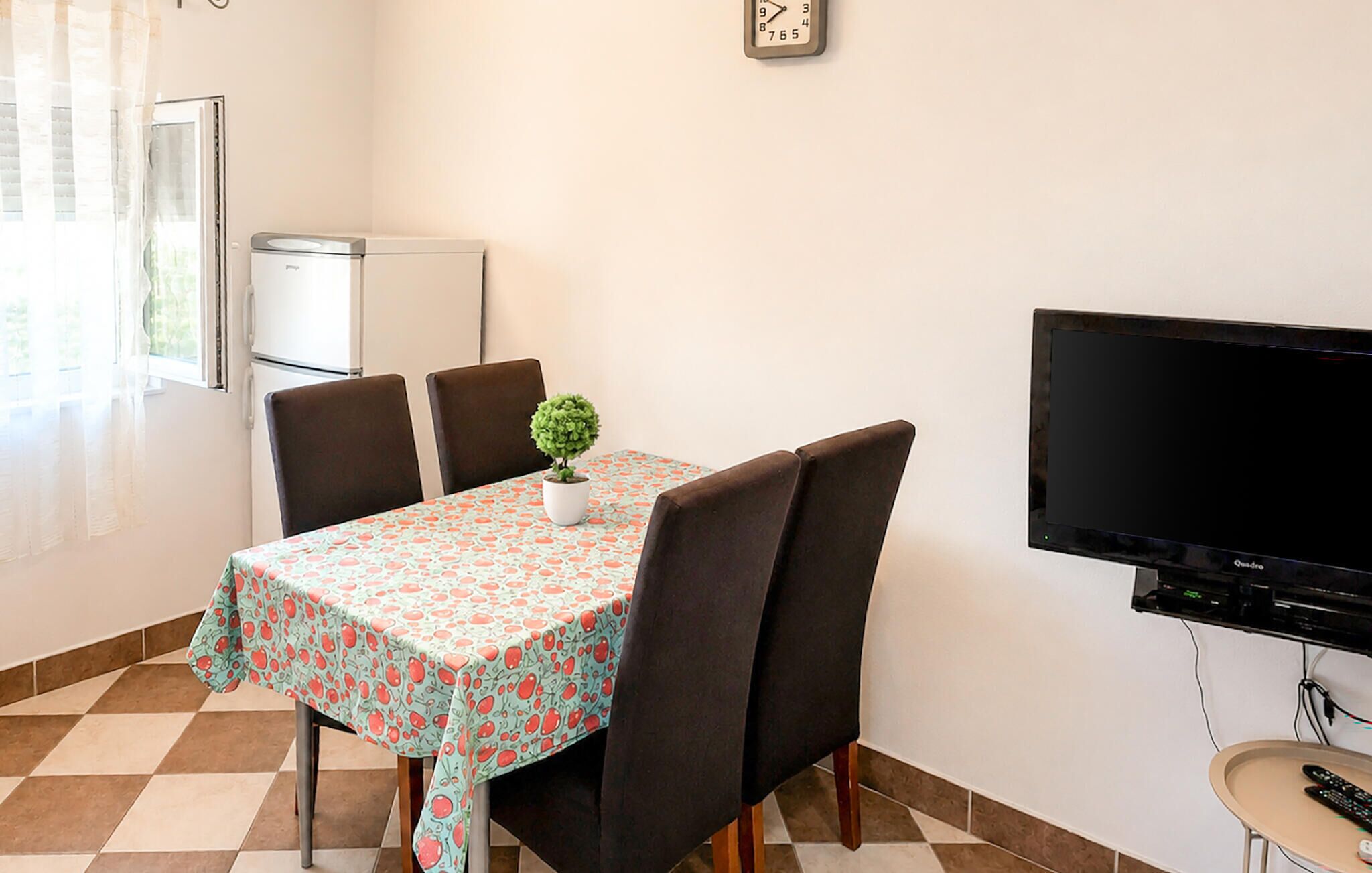 A dining area with a table, chairs, a small potted plant, a refrigerator, and a television.