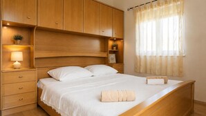 A bedroom with a wooden bed, a nightstand with a lamp, a shelf with a plant, and a window with sheer curtains. - Kakma Apartment private pool (Kakma)