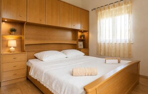 A bedroom with a wooden bed, a nightstand with a lamp, a shelf with a plant, and a window with sheer curtains. - Kakma Apartment private pool (Kakma)
