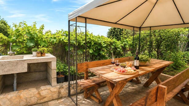An outdoor patio with a wooden table, benches, a grill, and a large umbrella. - Kakma Apartment private pool (Kakma)