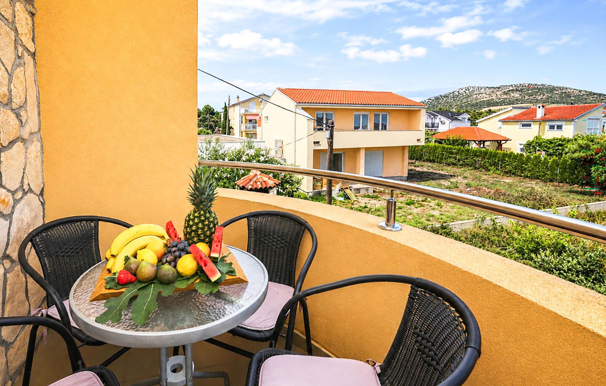 A balcony with a table set with fruit, wicker chairs, and a view of houses and greenery.
