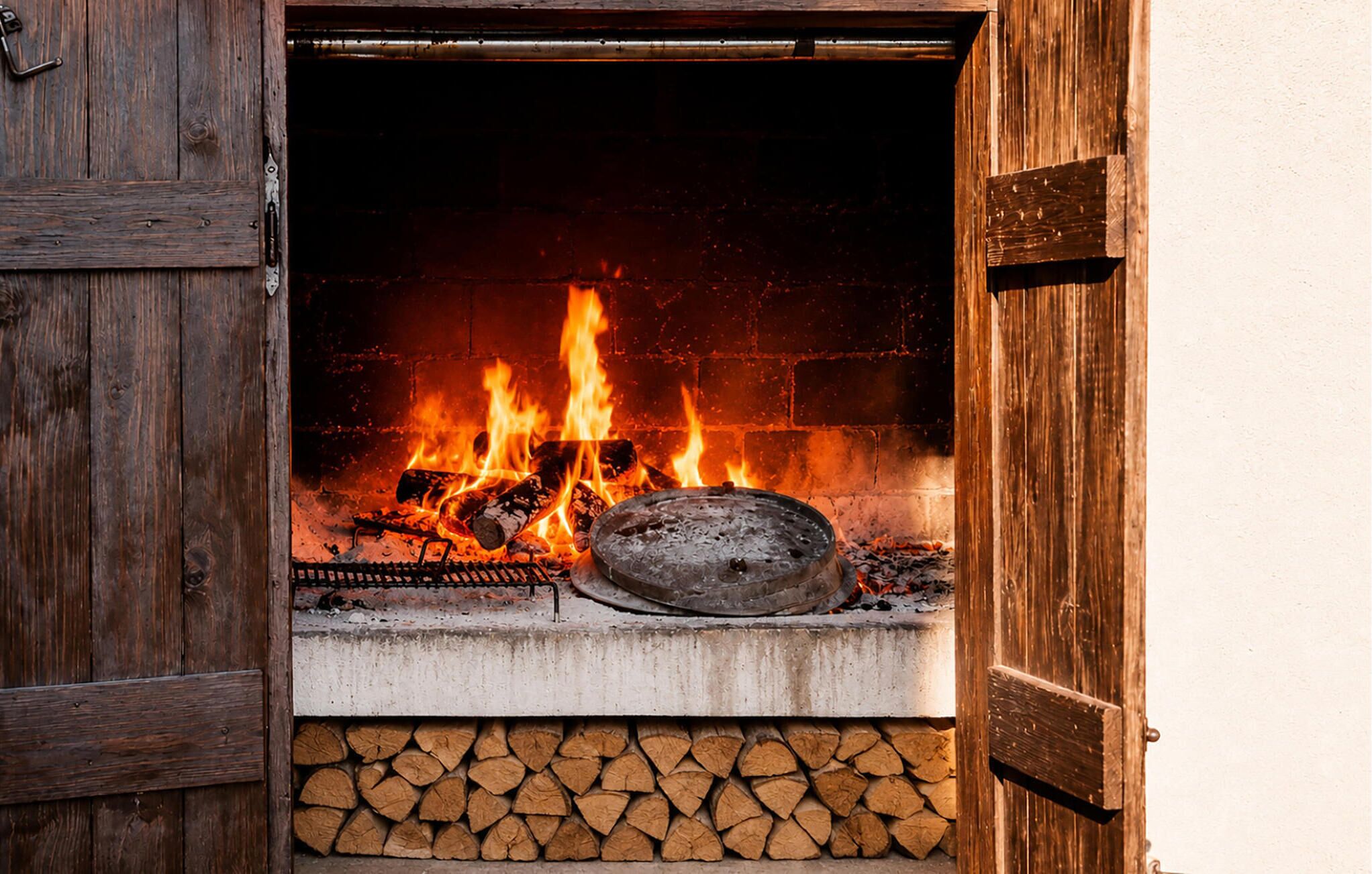 A fireplace with burning logs, a metal grate, and a stack of firewood.