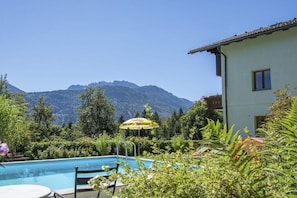 Una piscina con montañas al fondo, un edificio con balcón y abundante vegetación.