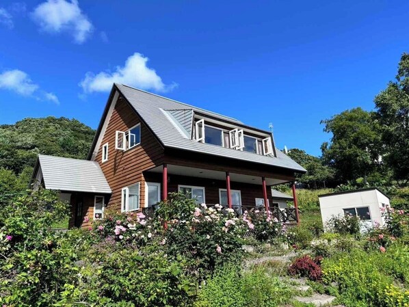 Une maison à deux étages, dotée d’un toit en pente, d’un porche avec des piliers rouges et d’un jardin où poussent diverses plantes et fleurs.