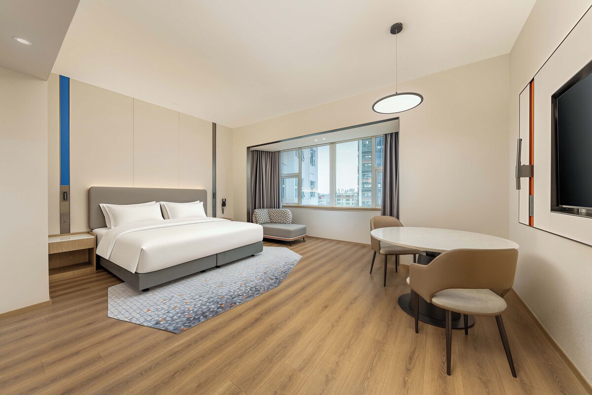 Habitación de hotel moderna con cama, escritorio, silla, mesa de comedor y un amplio ventanal con vistas a la ciudad.