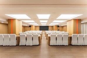 A conference room with rows of tables and chairs, a large screen at the front, and a high ceiling with skylights.