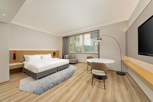 Una habitación de hotel moderna con cama, mesita de noche, silla, televisor y un amplio ventanal.