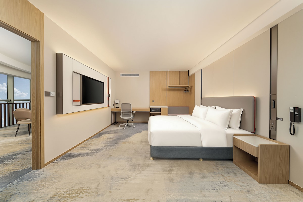 Habitación de hotel moderna con una cama grande, un escritorio, una silla, un televisor y vistas al balcón.