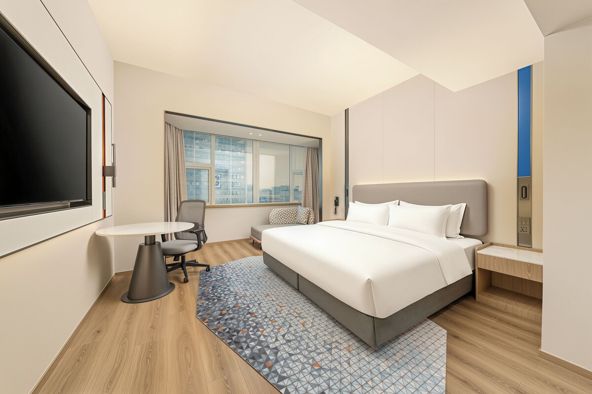 Habitación de hotel con cama, escritorio, silla, televisión y ventana con vista a la ciudad.
