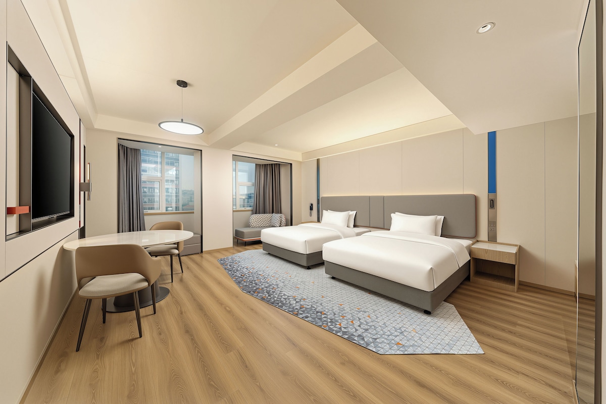 Habitación de hotel con dos camas, un escritorio, una silla, un televisor y un ventanal grande.