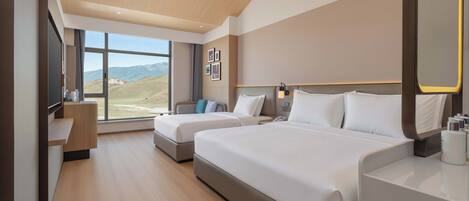 A hotel room with two beds, a desk, a TV, and a large window with a mountain view.