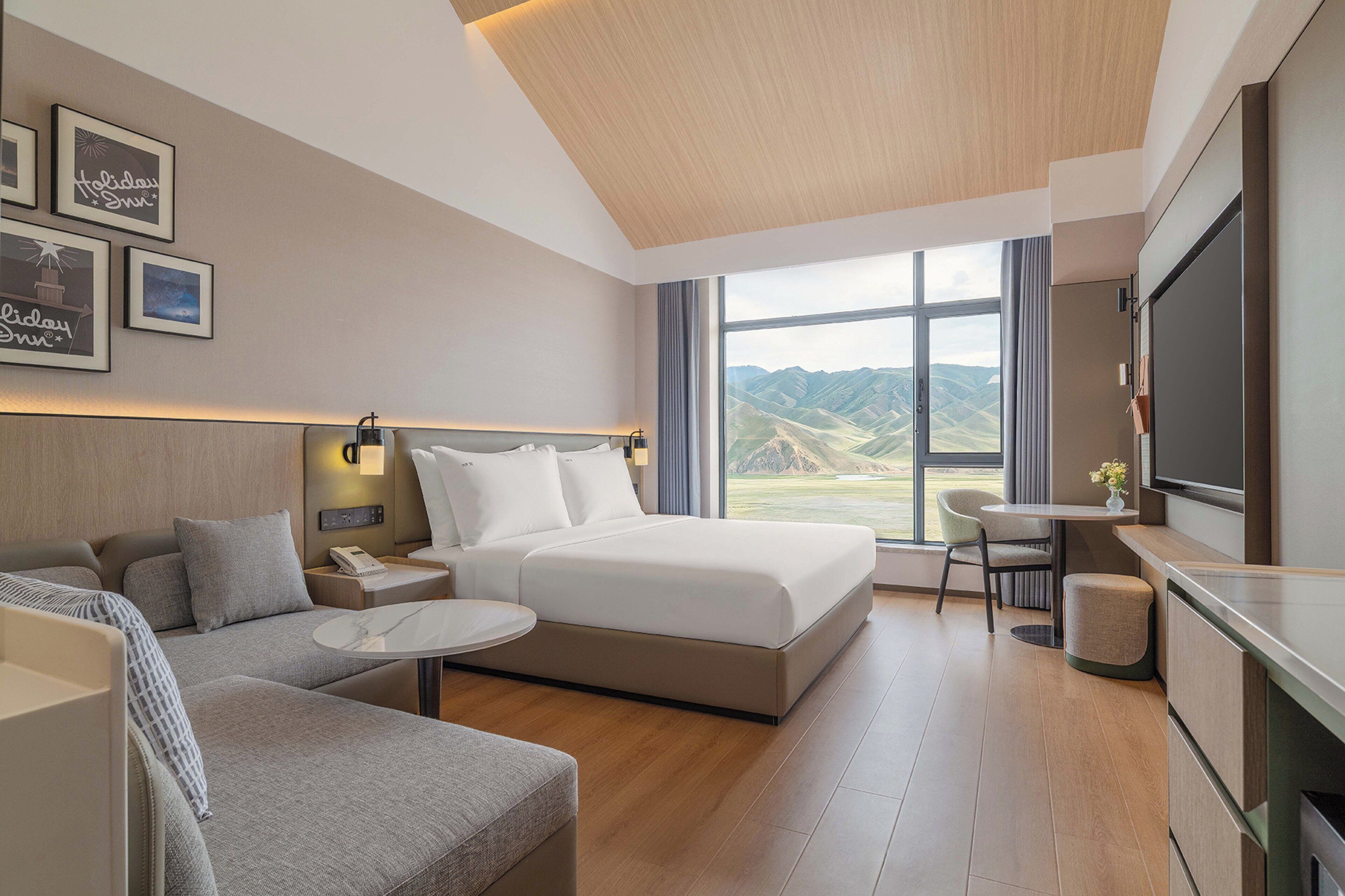 A modern hotel room with a bed, sofa, TV, and a view of mountains.