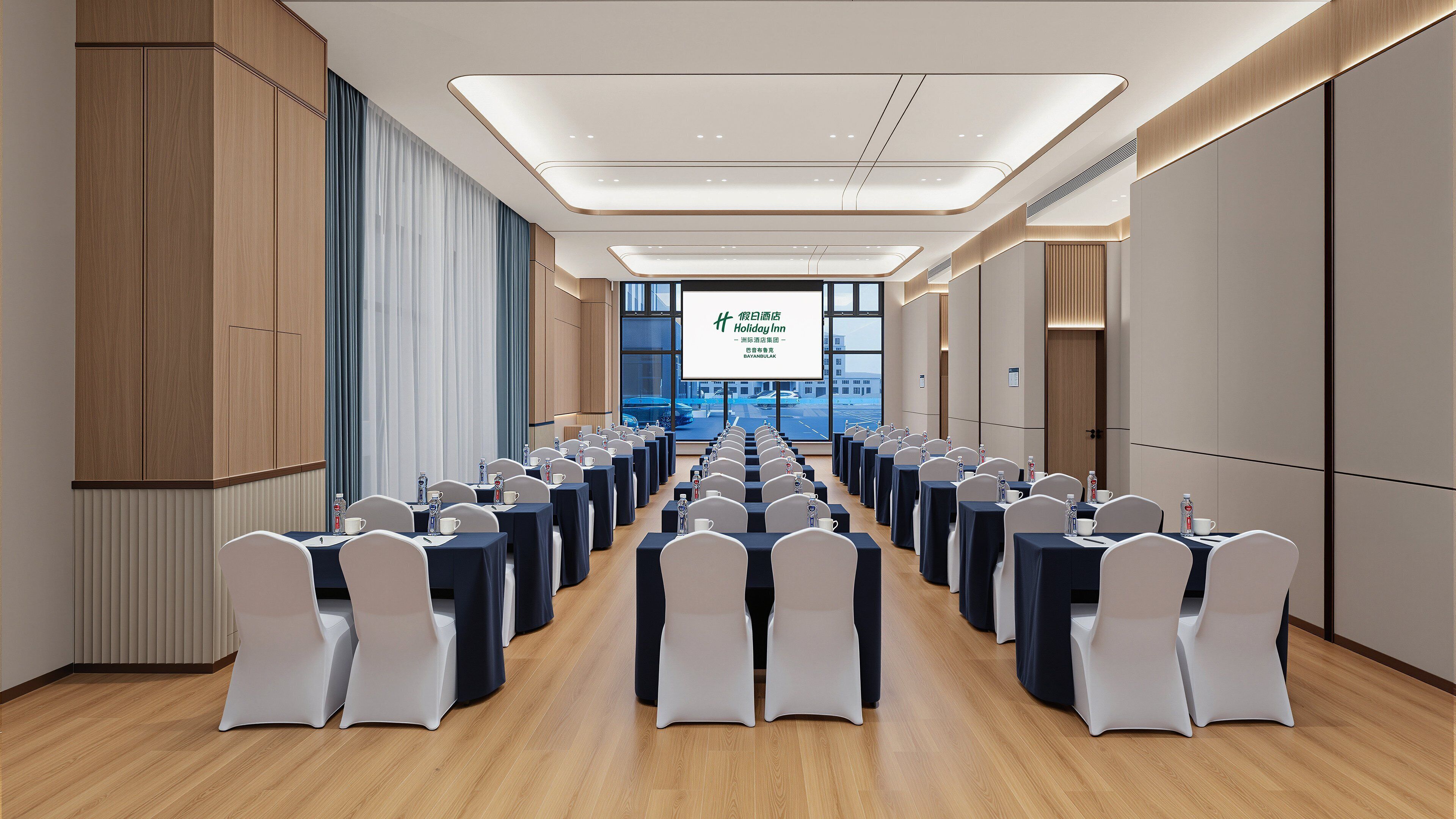 A conference room with rows of tables and chairs, a large screen displaying text, and a view of the outside through large windows.